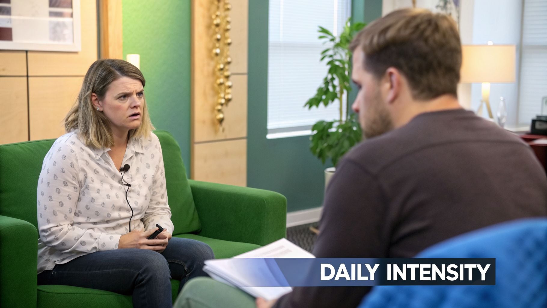 A woman with a microphone clip sits on a green couch, looking concerned while speaking to a man.