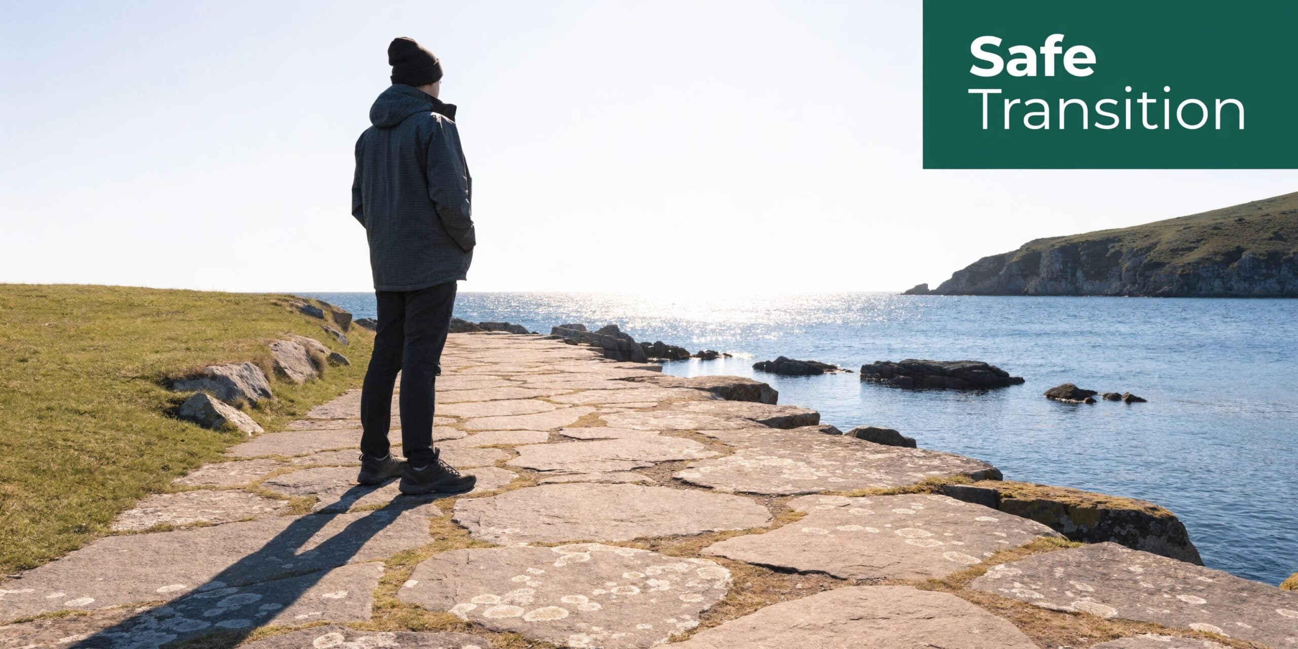 A person wearing a jacket and hat stands on a stone path looking out at the ocean.
