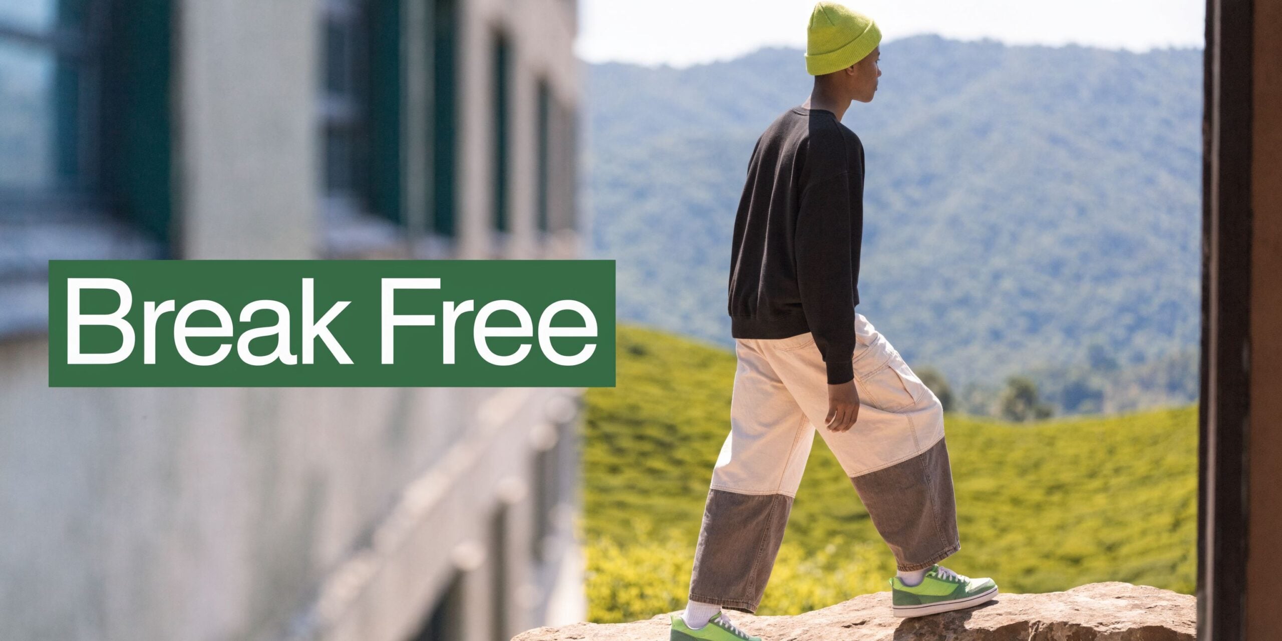 A person wearing a bright green beanie, black sweater, and two-toned pants walking along a ledge outdoors.