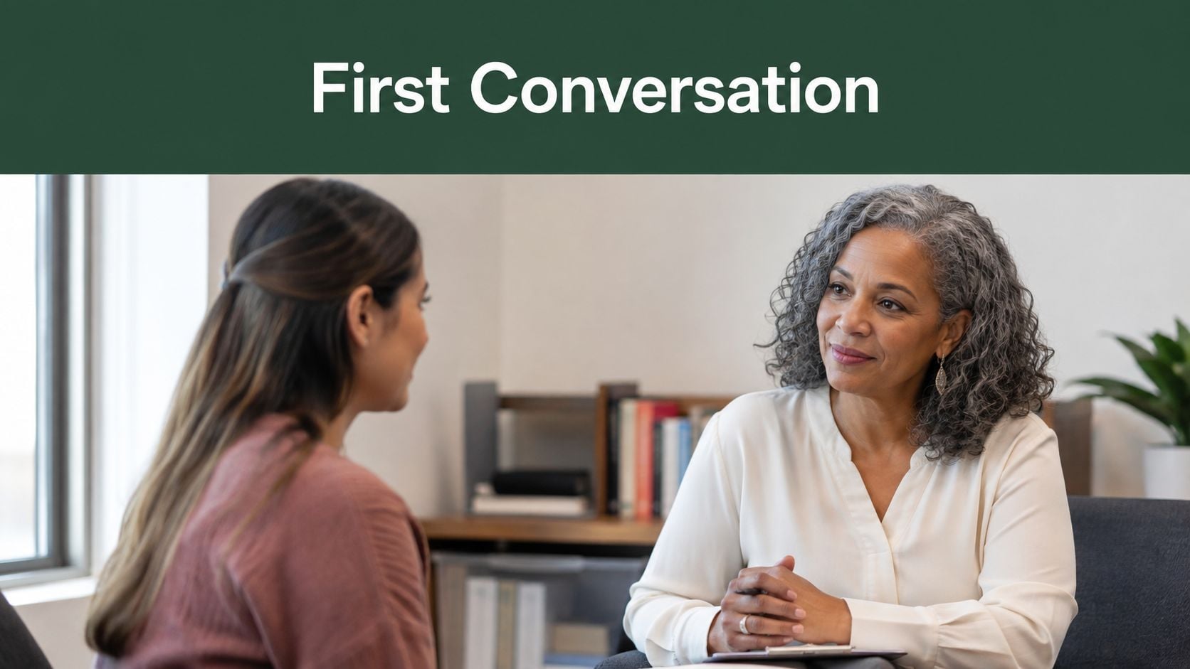 A professional woman with curly gray hair listens attentively to a young woman during a counseling session.